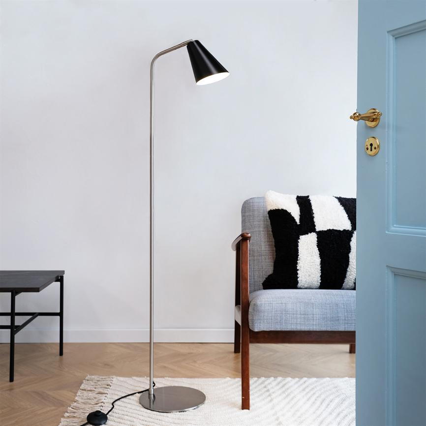 Floor lamp stands and casts warm light downwards by a gray sofa with a black and white pillow; open light blue door, small black side table, light carpet and wooden floor in a quiet corner.