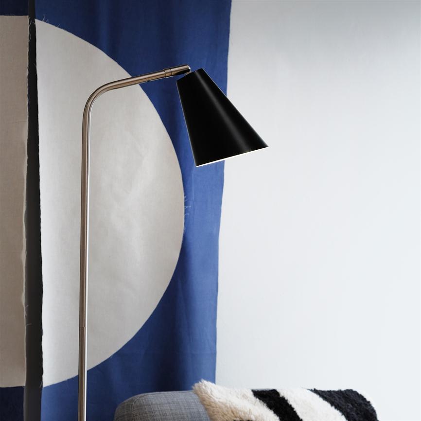 Floor lamp with black cone shade bends over a gray sofa; it shines down in front of a blue-and-white curtain with a large circle motif, by a black-and-white pillow.