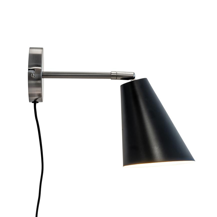 Black conical wall lamp points obliquely downwards and casts light; mounted on a brushed metal arm with round wall bracket and visible black cord against white background.