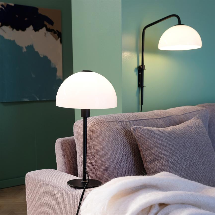 A white dome table lamp stands and shines on the armrest of a beige sofa; a corresponding wall lamp casts light against a mint green wall with an abstract painting in a corner of the living room.