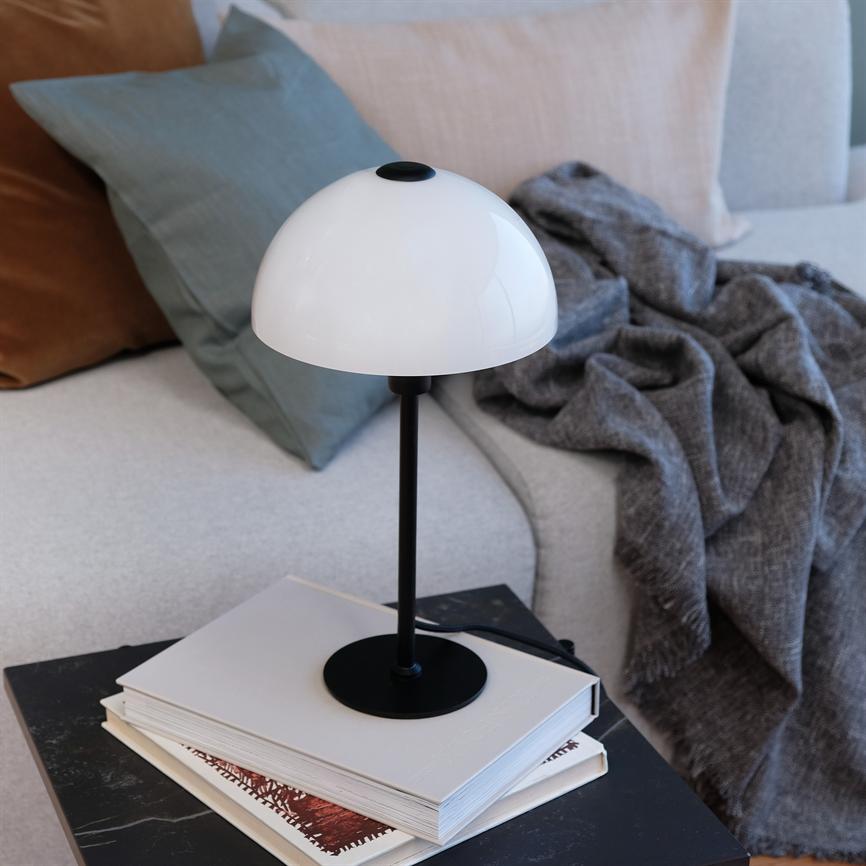Table lamp with white dome on black rod rests on top of a stack of books on a small black table; next to a light sofa with colored cushions and a gray rug.