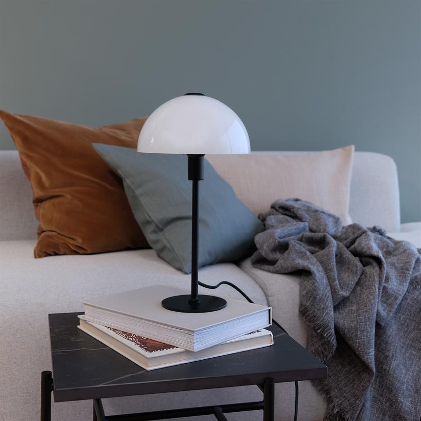 A table lamp stands on a stack of books on a small table; in the background a light sofa with brown and green cushions and a gray crumpled carpet in front of a blue-grey wall.