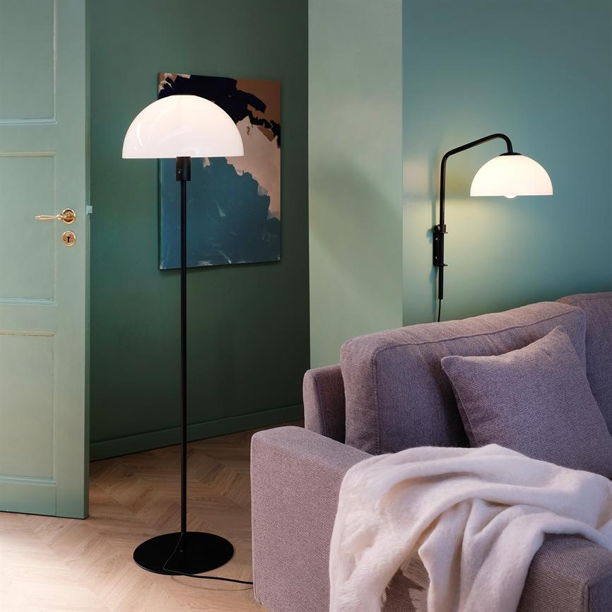 A black floor lamp with a hemispherical white shade glows softly; a matching wall lamp above a light gray carpeted sofa, set against green walls and an abstract painting.