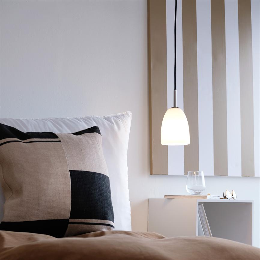 Lamp hangs and shines above a bedside table; next to it, a large checkered pillow sits on a bed, and the bedside table holds a water glass, glasses and a striped wall decoration.