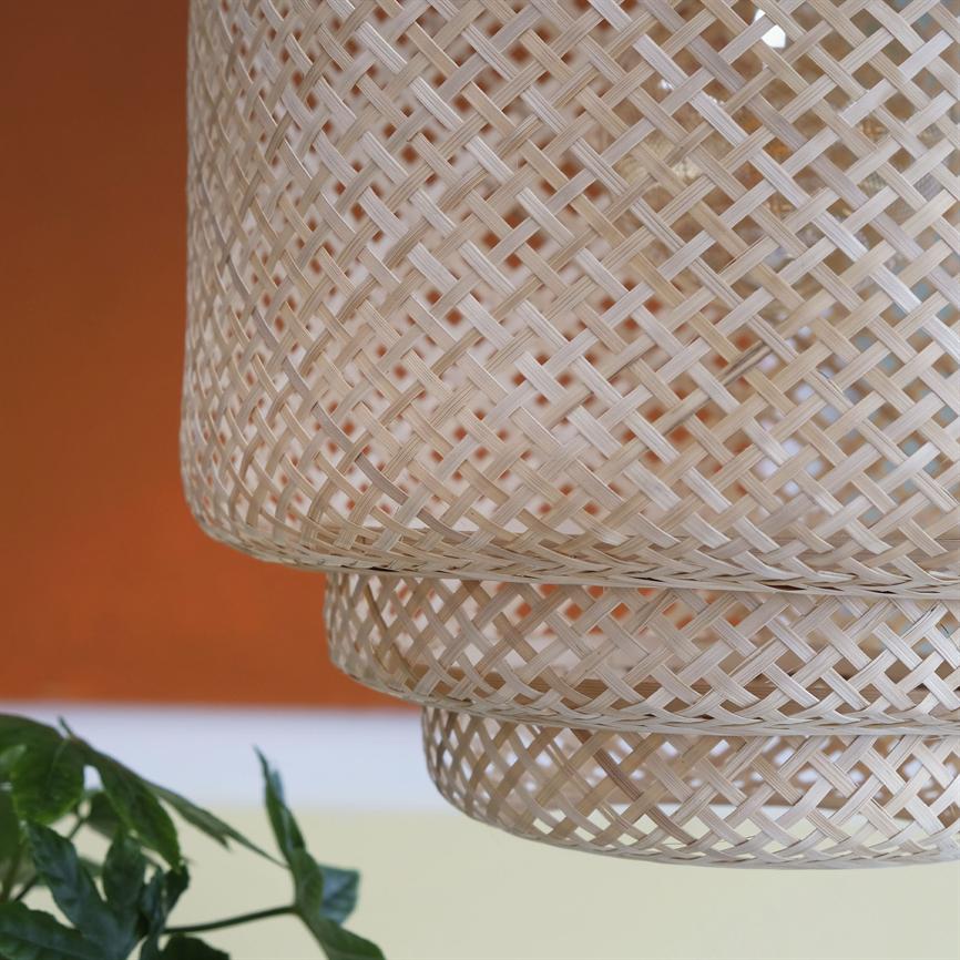 A wicker lampshade of bright bamboo hangs in the foreground in front of an orange wall; cross pattern is emphasized and green leaves poke into the lower left corner.