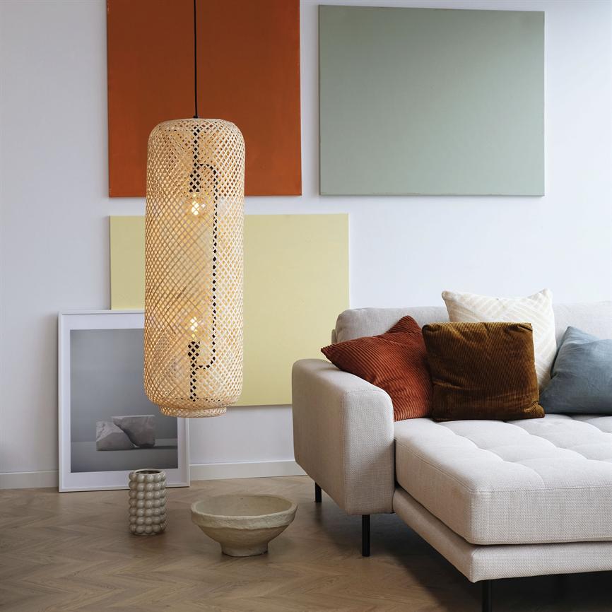 A wicker rattan pendant lamp hangs lit in front of colored wall panels; a light gray sofa with rust, brown and light blue cushions stands on the right in a bright corner of the living room with jars on the wooden floor.