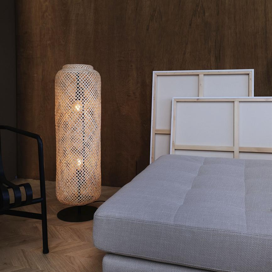 The woven rattan floor lamp is lit and casts warm light next to a light gray upholstered mattress, front-facing canvases and a dark wooden wall on a parquet floor.