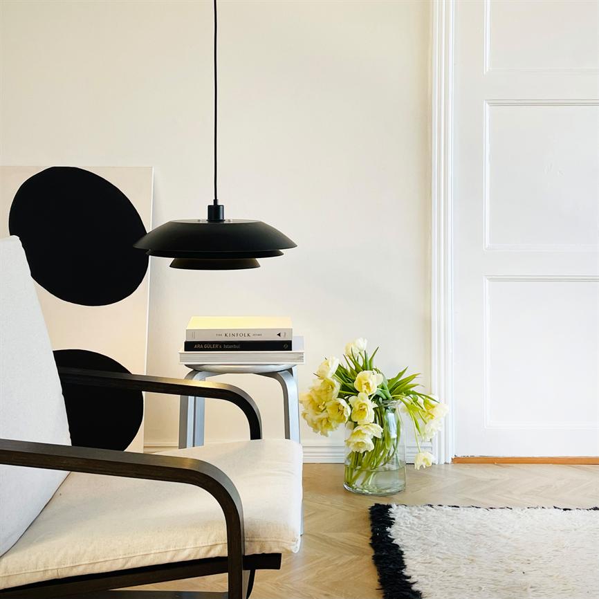 Black ceiling lamp hangs above a small side table with books in a bright, minimalist living room with a beige armchair, wooden floor, white door and a glass vase with yellow flowers.
 Text: THE KINFOLK TABLE; ARA YELLOWS - Istanbul