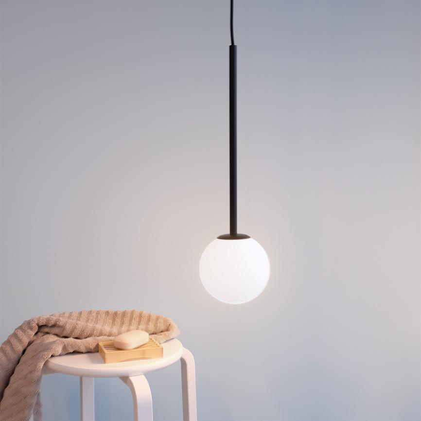 A round, white pendant glows softly; it hangs above a small white table, where a folded beige rug and a soap on a wooden tray lie in a bright, minimalist space.