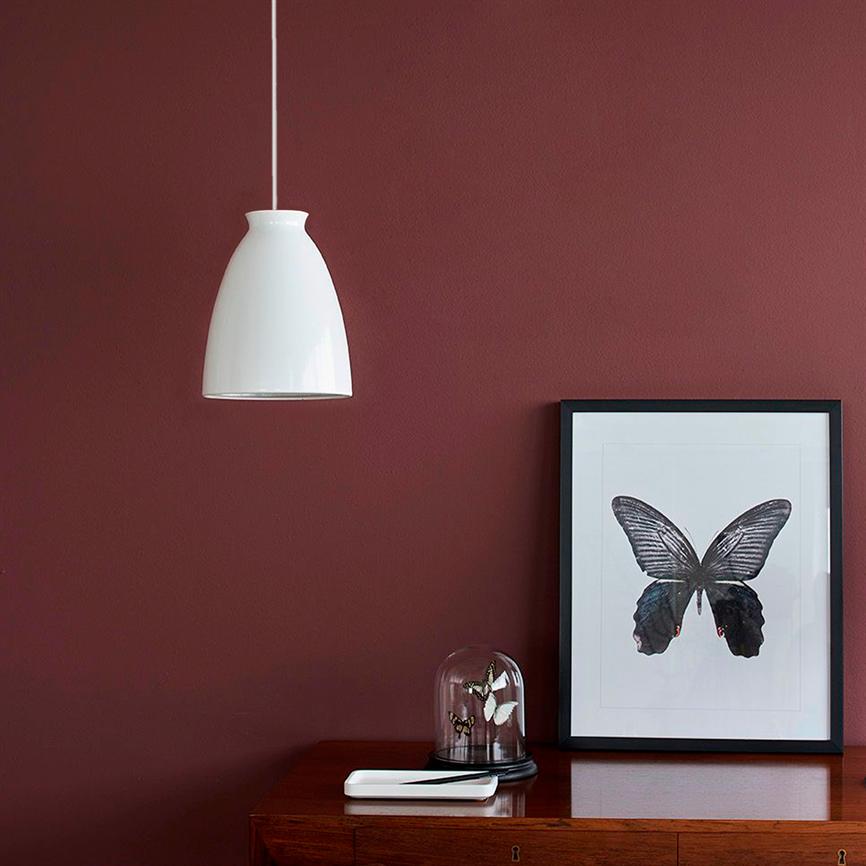 A white pendant lamp hangs above a wooden cabinet, where a framed butterfly print stands next to a glass bell with two small butterflies; the background is a dark red wall.