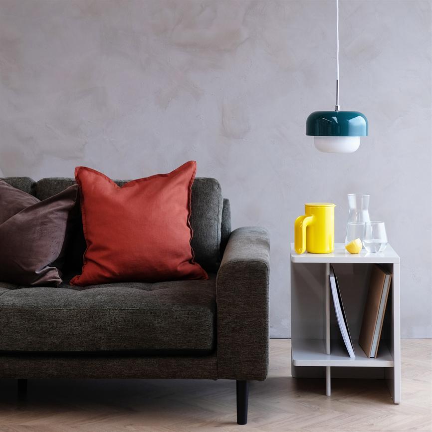 Gray sofa with a red decorative pillow, rests calmly on light wooden floor; next to it is a white side table with a yellow jug and glass, above it hangs a green ceiling lamp.