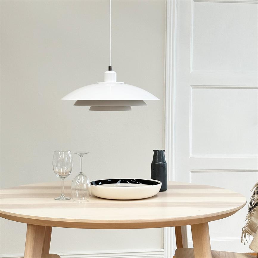 pendant hangs over a round light wooden table; on the table is a black and white bowl, a black decanter and two wine glasses (one upright, one upside down) in front of a white paneled door.