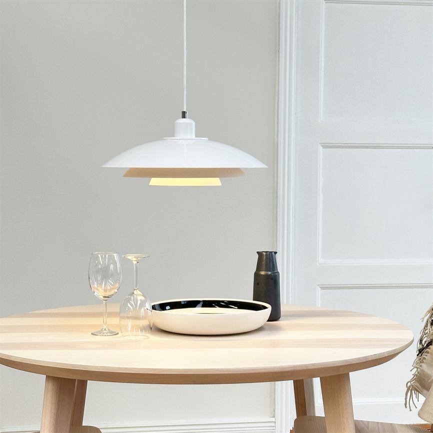 A white hanging lamp hangs above a round light wooden table, where a white bowl, two wine glasses (one upside down) and a dark jug stand in a bright, minimalist space.