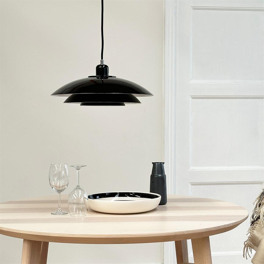 A black pendant lamp hangs above a round bright table; a black and white bowl, two wine glasses (one upright, one upside down) and a black jug stand in front of a white wall with a door.