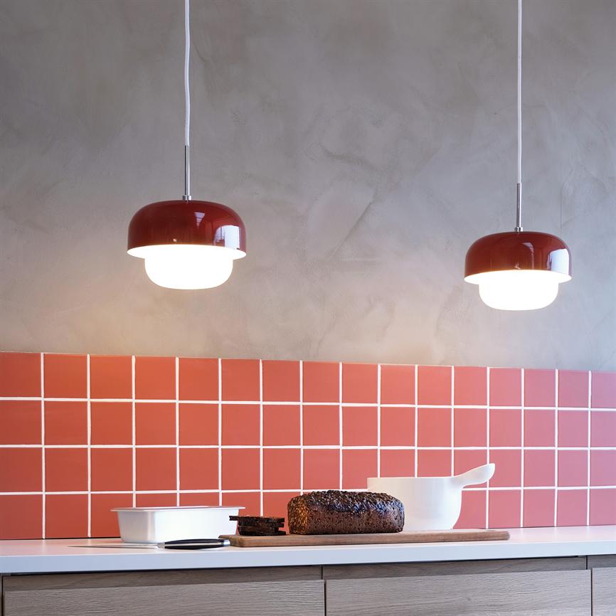 Two red pendant lights shine above a kitchen table; on the table is a cutting board with rye bread and slices, a white bowl and a white dish in front of a red tiled wall.