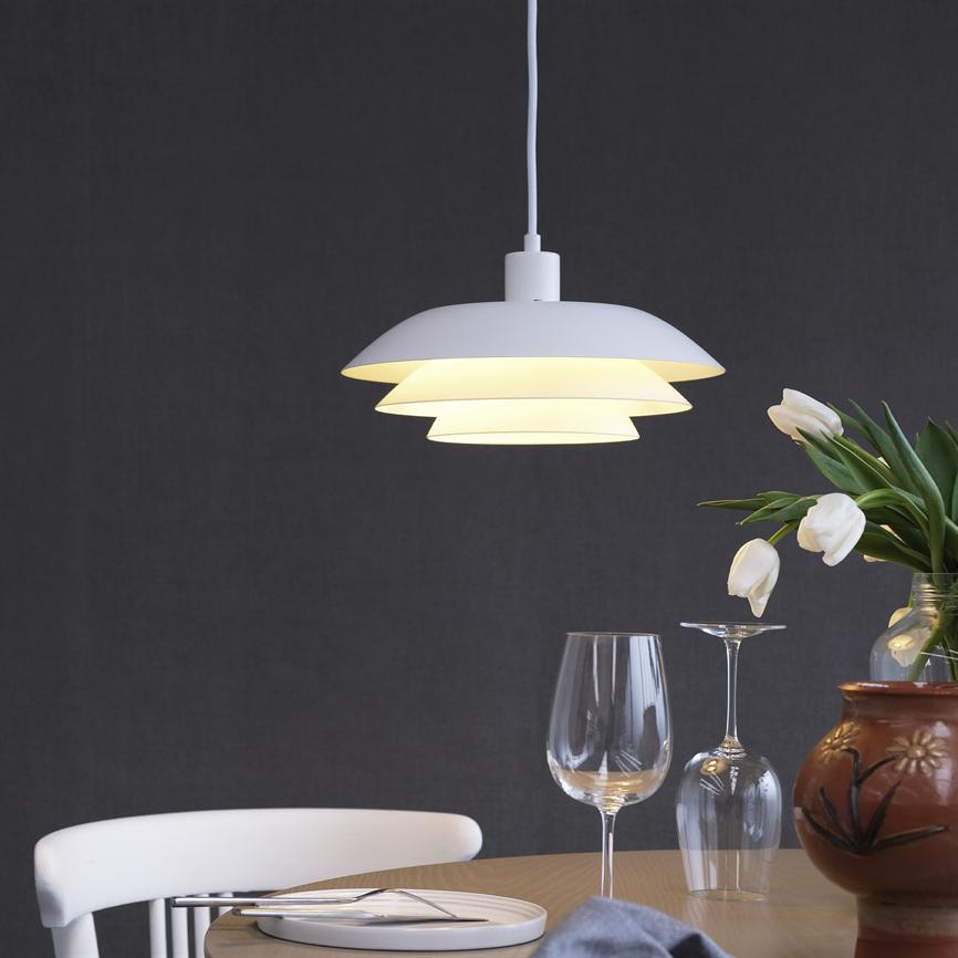 White pendant lamp hangs and shines above a round wooden dining table with plate, cutlery, two wine glasses, a white chair and a vase with white tulips in front of a dark gray wall.