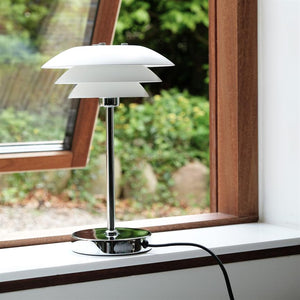 A white tiered table lamp stands on a white windowsill, connected by black cord; an open wooden window frames a blurred green garden outside.
