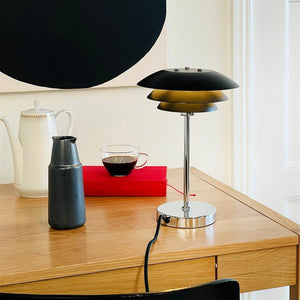 A black-metallic table lamp stands on an oak desk next to a glass cup of coffee, a black jug, a white jug and a red book in front of a white wall with a black circle.