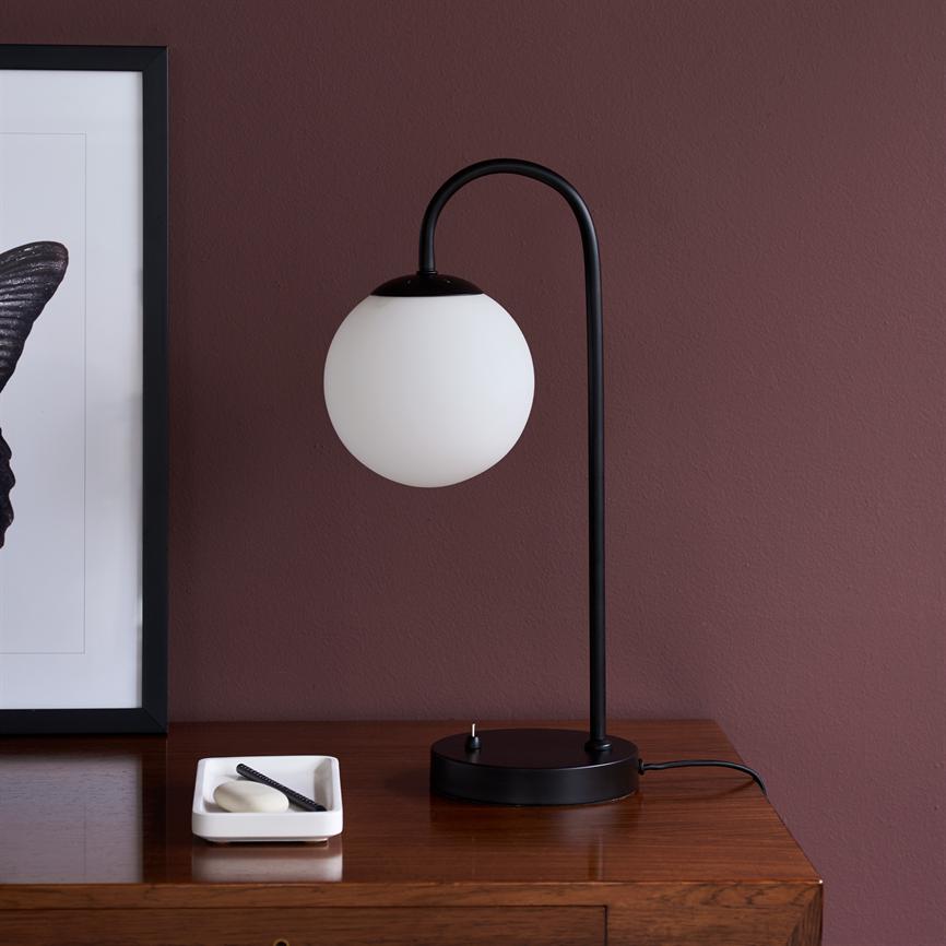 Black metal table lamp with white glass shade hangs from curved arm, stands on wooden drawer; beside a white tray of soap and comb; background: bordeaux wall and framed poster.