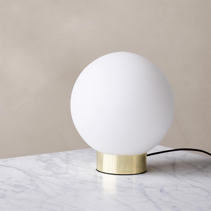 White, round lamp rests on a low cylindrical brass base; it stands without a light on on a white marble tabletop, with a black cord trailing behind, in front of a neutral beige wall.