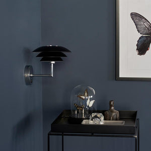 Black wall lamp mounted on a dark blue wall; on a small black table stands a glass dome with three mounted butterflies and a rustic ceramic figure next to it.