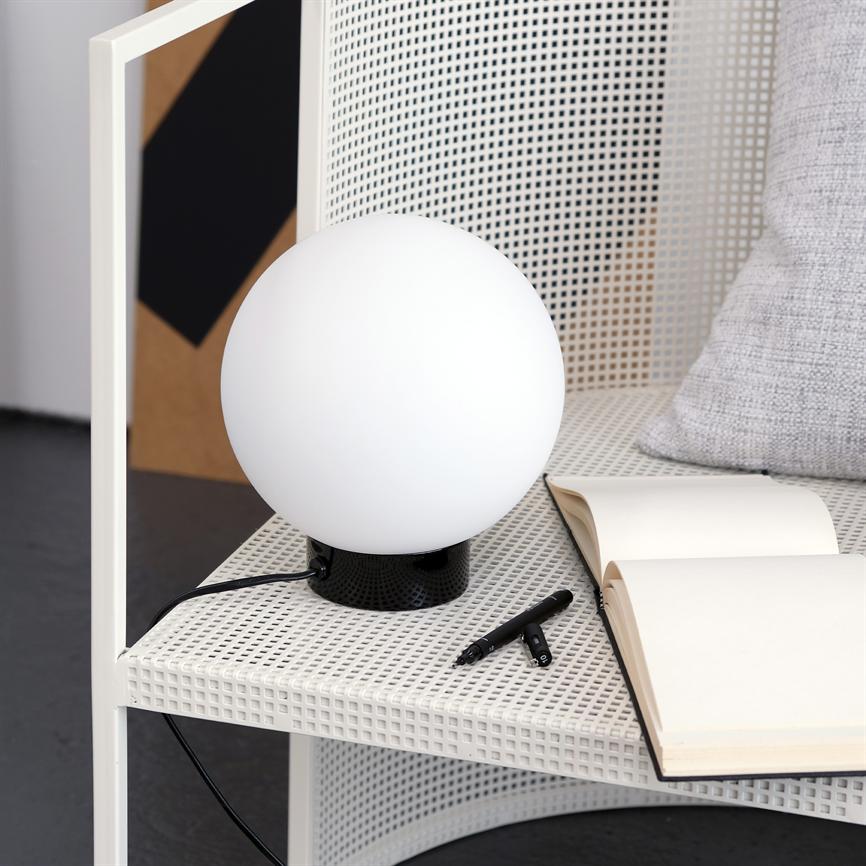 A round white lamp stands on a white perforated shelf, glows dimly; next to it is an open notebook and two black pens, with a gray pillow in the background.