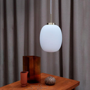 A white oval pendant hangs over a wooden table; on the table is a tall brown glass vase, a small terracotta cup and a twine key with a loose string; the background is gray curtains.