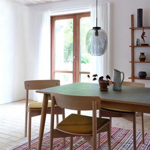 A wooden dining table with a green table top stands centrally, carrying a clay pot with a plant and a white jug; surrounded by wooden chairs, a hanging glass pendant, a window with a view and shelves on the wall.