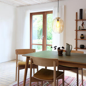 The dining table with four wooden chairs stands under a large hanging glass lamp; a jug and a small potted plant are on the table; large windows open onto a green garden section in a bright room.