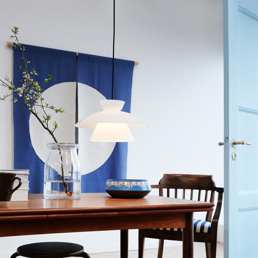 A white one pendant hangs over a wooden dining table; on the table is a glass vase with a flowering sprig and a blue patterned bowl, in front of a blue tapestry with a white circle and an open light blue door.