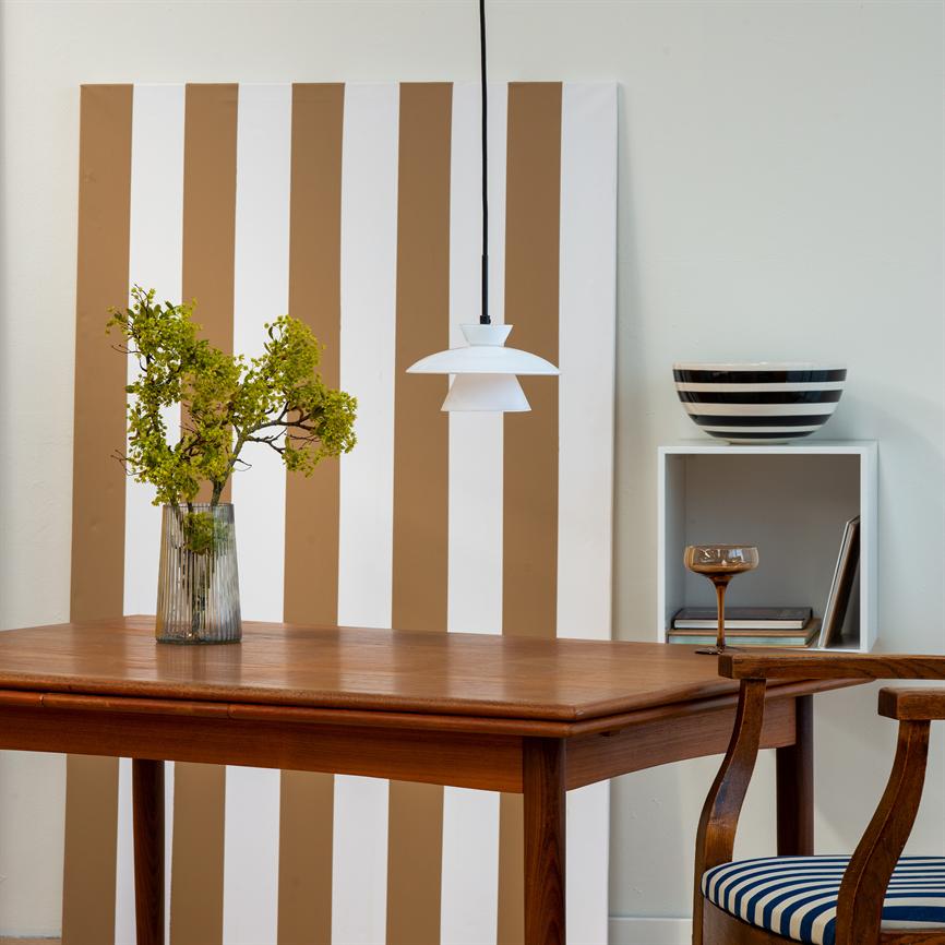 A wooden table bears a glass vase with green branches; above it hangs a white one pendant, in front a vertical brown-white striped plate; to the right shelf with bowl and chair with striped seat.