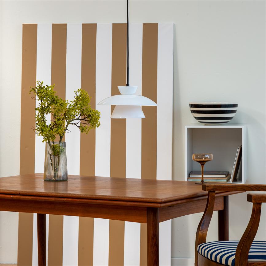 Wooden table stands in the foreground with a glass vase with green branches; a white one pendant hangs above the table, in front of a brown and white striped canvas and a wall shelf with bowl and chair.