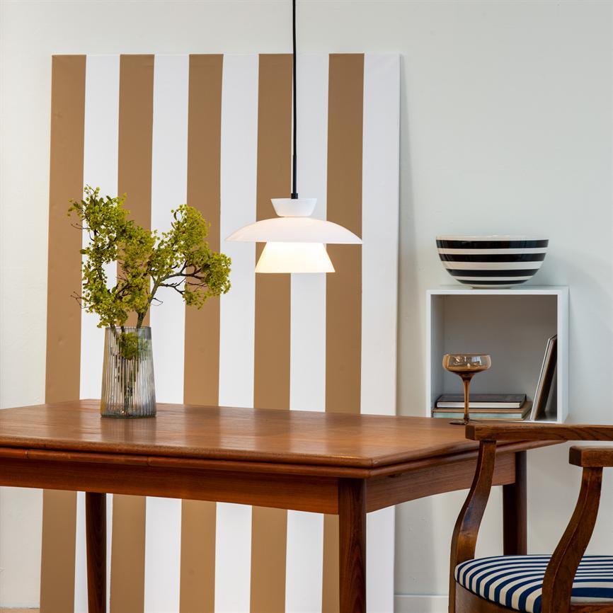 A white one pendant-lamp hangs above a wooden table and casts warm light; behind it is a tall white-and-brown striped plate, a glass vase with greenery and a small shelf with a bowl.
