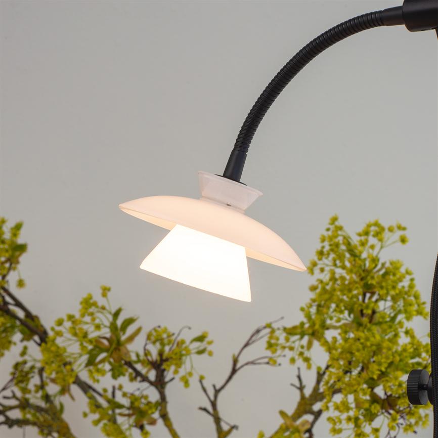 A white two-layer lampshade on a black flexible arm shines downwards; it casts warm light in front of a light wall with green, flowering branches in the background.
