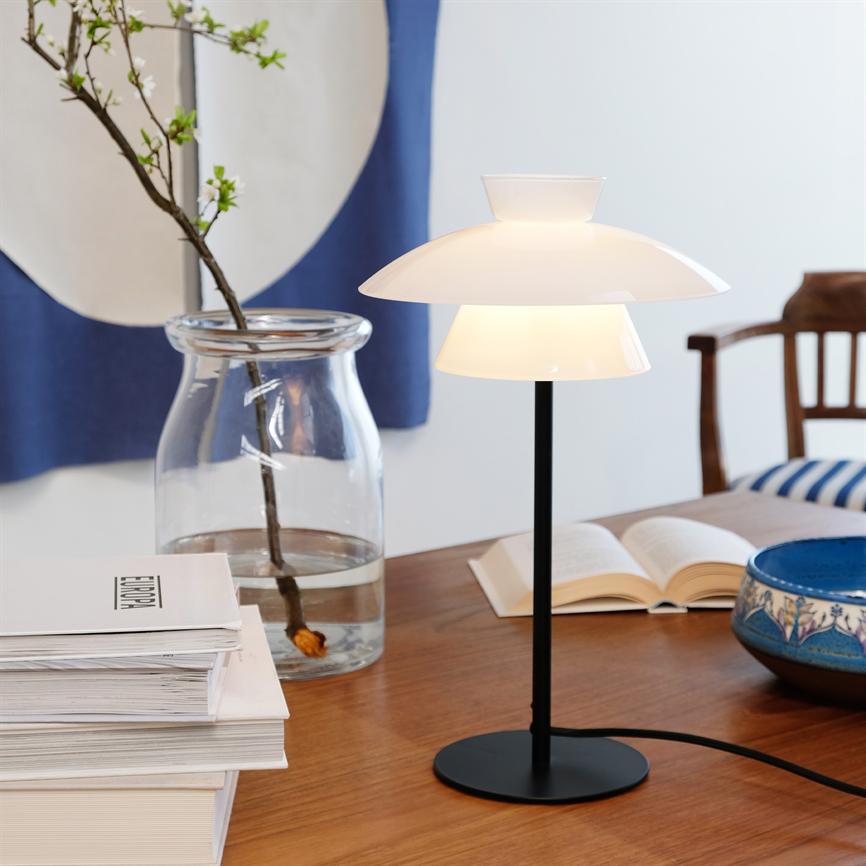 Table lamp glows softly; standing on wooden table by open book, blue ceramic bowl, glass vase with sprig and stack of magazines marked "EUROPA"; background: blue textile and wooden chair.