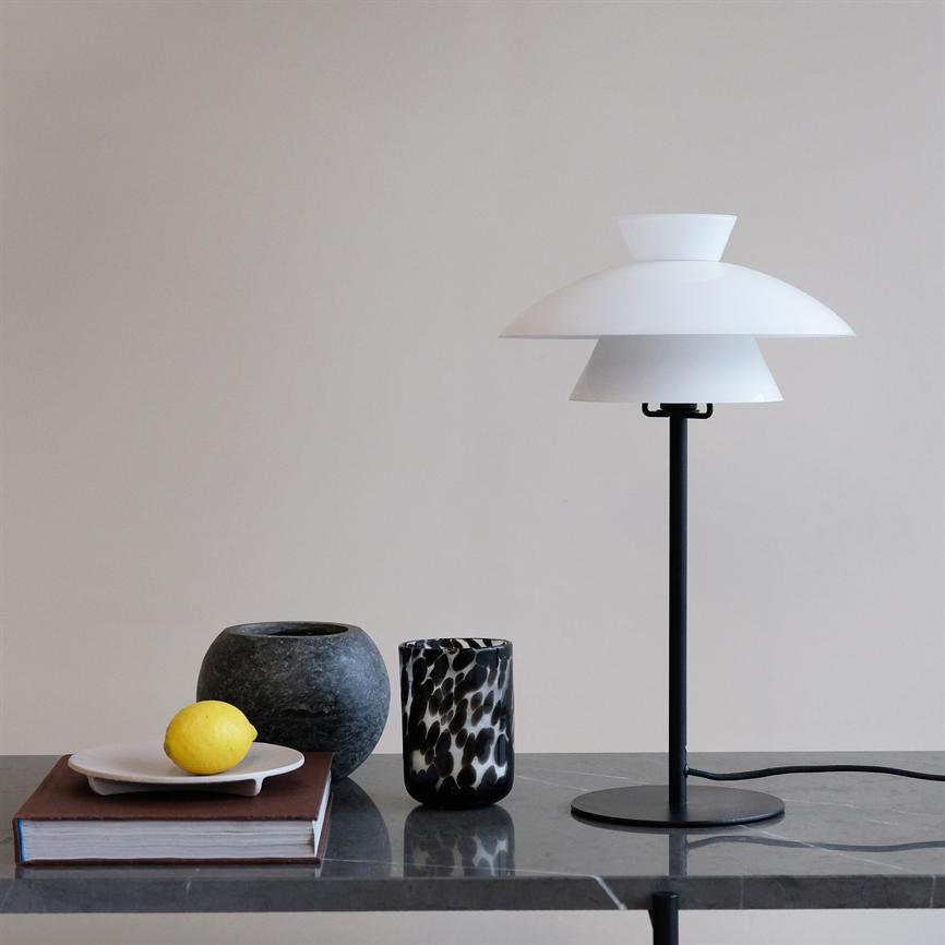 White, layered table lamp stands on a black base; next to it is a bound book with a white plate and a lemon, as well as two decorative jars on a gray marble table top.