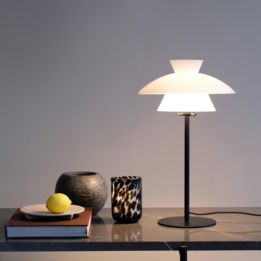 Table lamp emits soft, warm light over a marble table; next to it is a book with a plate and lemon, a round stone vase and a black and white patterned glass.