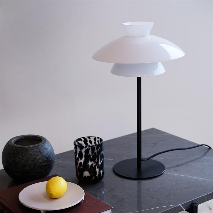A white, modern table lamp stands on a dark marble table; next to it is a yellow lemon on a small plate, a patterned glass and a round stone bowl.