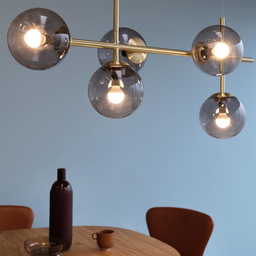 pendant with five smoky glass lamps, lit bulbs emitting warm light, hanging above a round wooden table with a dark bottle, small cup and brown chairs in front of a pale blue wall.