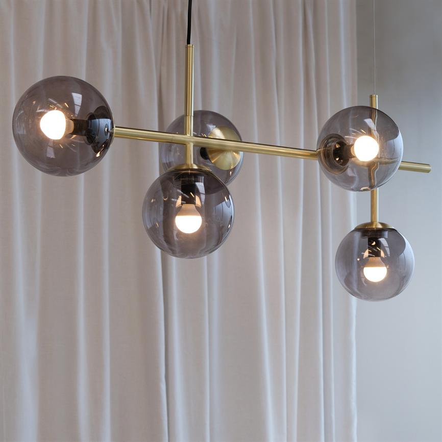 A modern ceiling lamp hangs with five smoky glass balls, some with lit bulbs; a brass rod connects the spheres — in front of soft white curtains in a bright room.