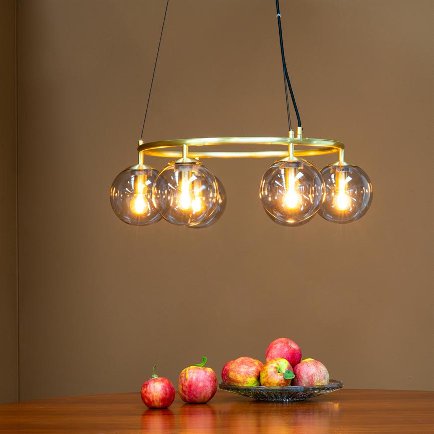 A ceiling lamp hangs and emits warm light: a gold ring with four clear glass balls shines above a wooden table, where a glass bowl with several red apples and two apples stands.