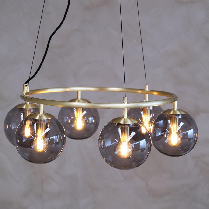 Gold ring with five smoky glass spheres, each containing lighted bulbs, emitting warm glow; the fixture hangs from black wires in front of a softly structured gray wall.