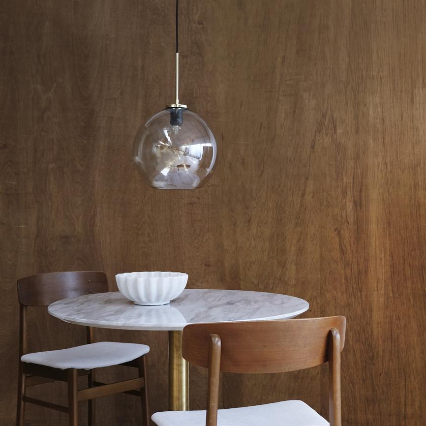 A glass pendant hangs above a round marble table with a white bowl, flanked by two wooden chairs in front of a warm, wood-clad wall.