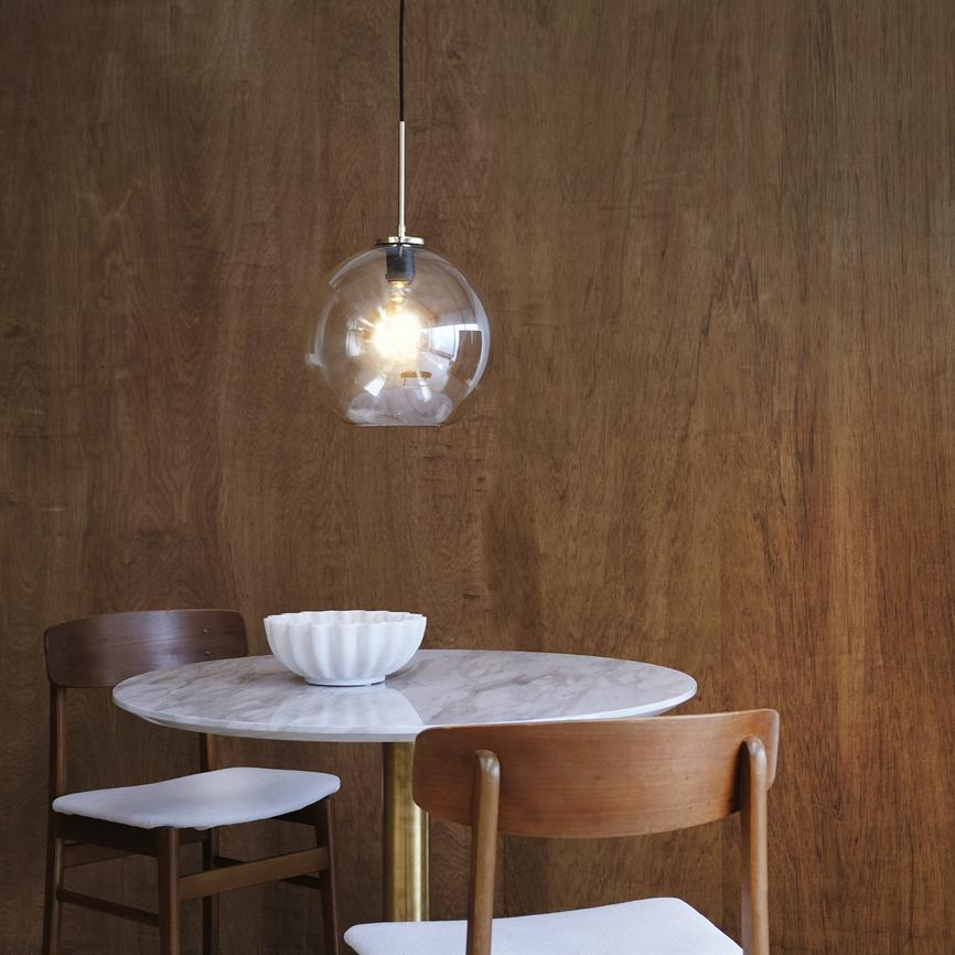 A hanging glass pendant lights warmly above a round marble table with a white bowl; two wooden chairs stand around the table in front of a dark wooden wall.
