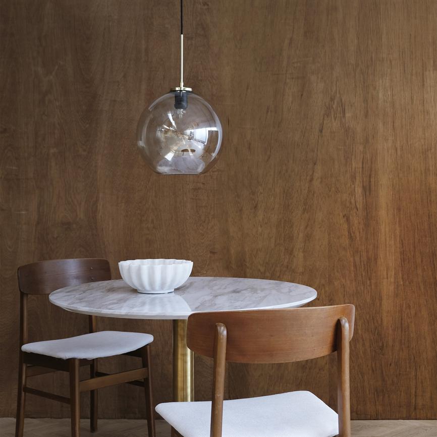 A round marble table stands with a white bowl; two tufted chairs with white cushions flank it, and a clear glass pendant hangs above the table in front of a warm wood-clad wall.