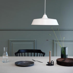 White pendant hangs over a set table; a black chair behind, glass carafe, a plate and fork, candle holder with candles and vase with tall flowers in front of green wall.