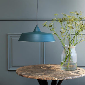 A blue one pendant hangs over a round marble table; a glass vase with delicate white flowers stands on the table in front of a grey-blue wall with framed panels in a calm, minimalist interior.