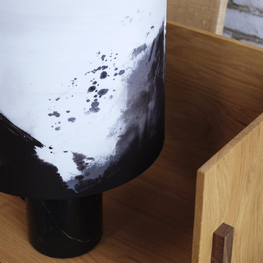 Lamp with black-and-white speckled shade stands on a black marble base inside a light wooden tray on a wooden table, close to a raw brick wall.