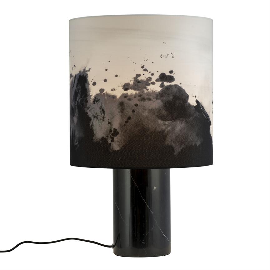 Table lamp with cylindrical black marble base and white shade, which has black ink or watercolor spots; stands on a white background, black wire lies bent in front.