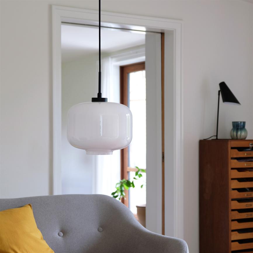 A white, round glass pendant hangs in the middle; in the foreground a gray sofa with a yellow cushion, in the background a wooden chest of drawers with a table lamp, a doorway and a green plant by the window.